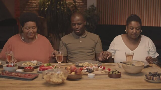 Medium close-up of African-American man and women sitting at table in dining room at home, holding hands and praying before meal