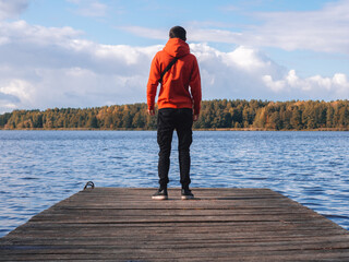 A young man in a bright orange hoodie on a wooden pier against the background of the lake. Autumn time, travel and active recreation concept.