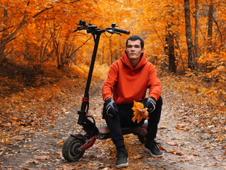 A young handsome man in an orange hoodie rides an electric scooter in an autumn park. Colorful seasonal portrait, forest road.