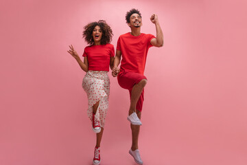 Full portrait of two young, energetic dark-skinned youths jumping on one leg against pink wall. Guy and girl with curly hair in red casual clothes. People sincere emotions lifestyle concep