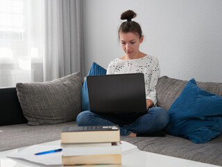 A young woman is seated on the couch and is using a laptop. There are books and notebooks on the table. Home office. E-learning.