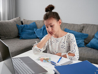 Young worried woman doing financial balance. The girl is counting the bills. There is money in euros and a laptop on the table. Summary of monthly costs. Rising living costs cause stress