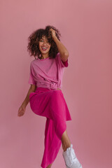 Slender American girl with dark afro hair dances to beat of drums with copy space. Model with natural makeup in trendy crimson pants and pink T-shirt. Lifestyle, female beauty concept