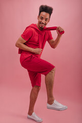 Full-length, inspired young man with afro hair lifted his leg up against pink wall. Stylish guy is looking at camera with pleasure, being joyful. Lifestyle leisure and beauty concept.