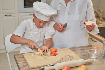 Mom and son in white chef's clothes cut pears with a knife in their home kitchen. An adult woman...
