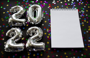 Inflatable numbers New Year 20212 on the left. Black background with shiny golden green and silver stars. White notebook, copy space