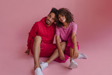 Full photo of young afro curly haired best friends sitting on floor in photo studio against pink background. Guy and girl are sitting face to face, looking at camera. Concept of enjoying moments.