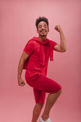 Young energetic afro guy in sports crimson suit with copy space is dancing on spot. Brunette with fluffy and curly hair carries positive energy, comedian posing against isolated background.