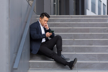 Asian businessman reads bad news from the phone, sitting near the office on the stairs, depressed, losing hope