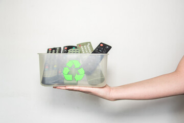 Una mano blanca sosteniendo una caja de plástico transparente con el símbolo del reciclaje que contiene controles remotos de tv sobre un fondo blanco. Mandos a distancia rotos y viejos - obrazy, fototapety, plakaty