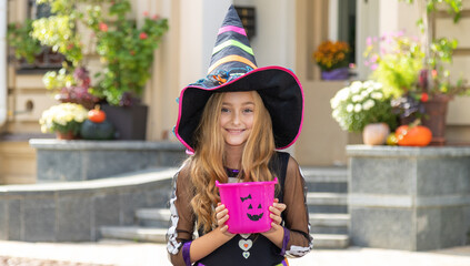 Little girl dressed as a witch with a bucket for sweets. Halloween holiday.