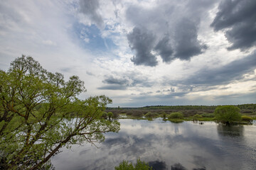 Landscape with a dramatic sky reflected in the river. Early spring, juicy May greens. Bright green foliage on trees and bushes.