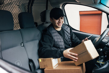 Smiling courier sitting on a driver's seat checking delivery information on mobile phone while holding box