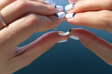 Fototapeta premium female hands with a gorgeous manicure with white nails and black splashes against the background of a bright blue sky and rocks with a wedding ring. Very beautiful female hands with gold rings