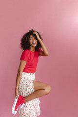 Young African woman stands half sideways and bends leg to knee on pink background. Softly touching curly and fluffy dark blond hair with thin hand. Looks beautiful in red T-shirt and skirt with slit.