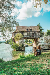 A Weimaraner breed dog next to a young girl sitting in front of the old mill on the medieval bridge in Vernon, Normandy - obrazy, fototapety, plakaty