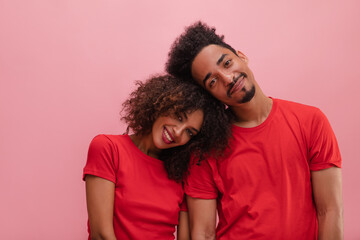 Fototapeta premium Pretty young mexican woman put her head on brother's shoulder, expressing happiness against pink background. Brunettes with curly hair, dressed in red t-shirts. Different emotions, leisure concept.