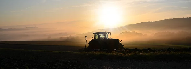 Muurstickers Tractor Trekker die het veld ploegt bij zonsopgang in de late zomer of herfst.