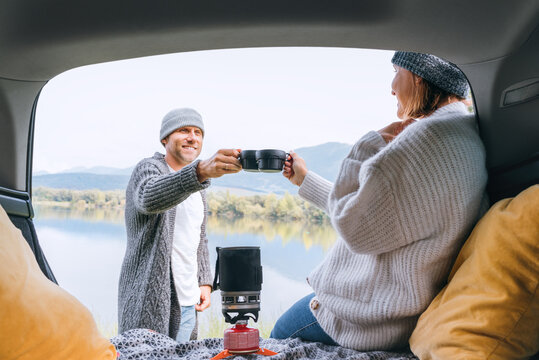 Car trunk view clinking tea mugs couple dressed warm knitted clothes enjoying gas stove prepared drink and mountain lake view. Cozy early autumn couple auto traveling concept image.