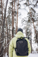 Fototapeta na wymiar Rear view of young man with backpack in yellow jacket walking in snowy pine forest in winter. People from behind. Local travel, exploring nature.