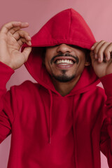 Close-up of handsome African man hiding eyes under hood over pink background. Smiling broadly with white teeth, wearing fashionable bright red sweatshirt. Spreading optimistic emotions and feelings.