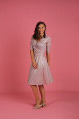 a young happy beautiful slender girl in a pink dress on a pink background looks at the camera