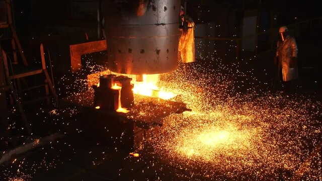 Laborer in protective suit stands by tank with liquid metal and bright sparks spread in contemporary steelmaking plant workshop