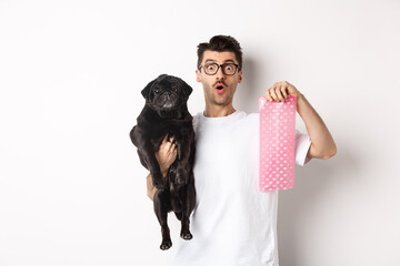 Image of hipster guy pet owner, holding cute black pug and dog poop bag, standing over white...