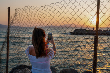 Back rear view of woman taking photo of sunset with mobile phone from behind fence