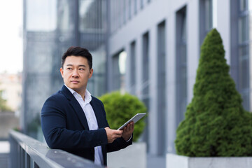 Fototapeta premium Serious asian boss reads news from tablet near modern office male businessman in business suit