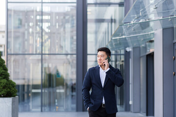 Fototapeta premium Successful Asian businessman businessman talking on a mobile phone while walking near the office of a large modern, serious and pensive employee