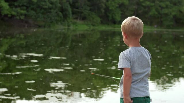 Back of a caucasian boy in t-shirt fishing at small pond in the park. A 4-years old boy with a fishing rod in calm summer evening. 