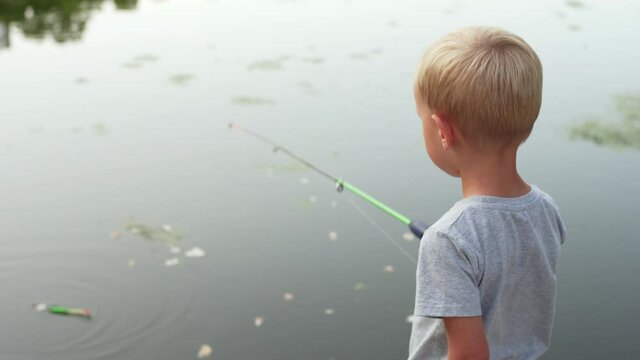 Back of a blonde caucasian boy in t-shirt fishing at small pond in the park. A 4-years old boy with a fishing rod in calm summer evening. 