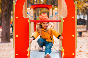 A little girl in the autumn plays on the playground in a yellow park jacket and jeans