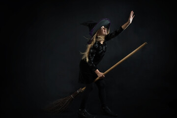 A little girl on a dark background in a witch costume and a wide-brimmed hat flies on a broom on Halloween.