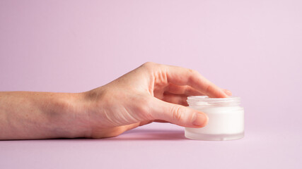 Hand holding cream jar. Hand holding face cream on purple background, close-up. Moisturizer in container. Side view