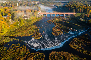 Venta Rapid waterfall, the widest waterfall in Europe and long red brick bridge in sunny, foggy autumn morning, Kuldiga, Latvia. Captured from above.
