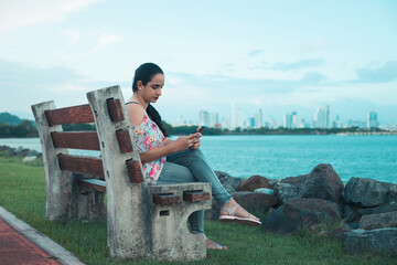Hispanic girl checking the cell phone, sitting on the bench near the sea