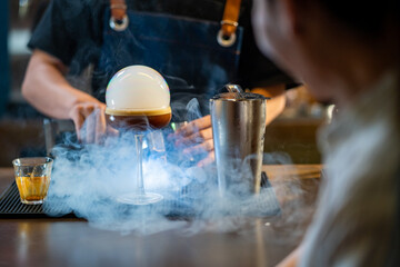 Asian man barista shaking iced black coffee in shaker and pouring in cocktail glass on bar counter at coffee shop. Male coffee shop owner serving cold drink coffee to customer. Small business concept.