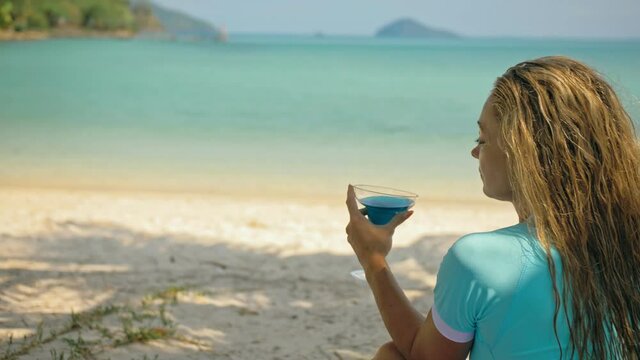 The woman is holding a glass of blue curacao cocktail, on beach azure blue sea of a tropical island. Blurred beautiful seaside background. Celebrate summer holiday concept of leisure, travel, relax.