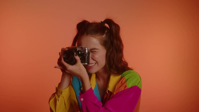 Lovely Smiling Young Woman In Two Ponytails Taking Photos With Mirrorless Camera Against Orange Background. medium shot