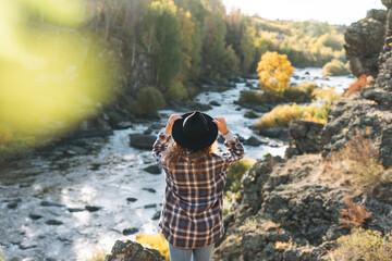 Young beautiful woman with curly hair in felt hat and plaid shirt in jeans looks at magic view of mountains and river, hiking on autumn nature