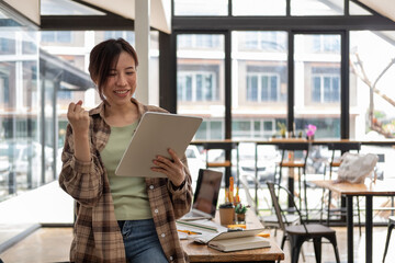 Fototapeta premium Young Asian woman celebrate success or happy pose during hand holding digital tablet, woman working at her office