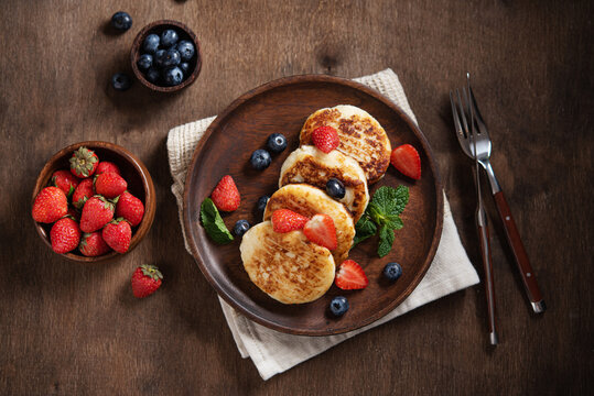 Cottage cheese pancakes or syrniki with blueberries, strawberries and fresh mint on a dark wooden background. The concept of a home-made healthy breakfast. Top view image