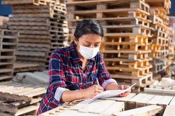 Woman manager in protective mask keeps records of building materials in the open area of a...