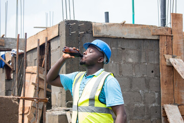 A thirsty male African construction worker drinking from a bottle on a building site and the man is also wearing a blue safety helmet known as hardhat and green reflective traffic jacket