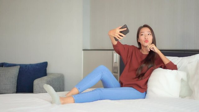 Smiling Asian woman takes selfies on smartphone camera while sitting on the bed at home