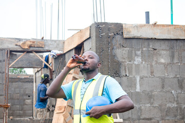 A thirsty male African construction worker drinking from a bottle on a building site and the man is...