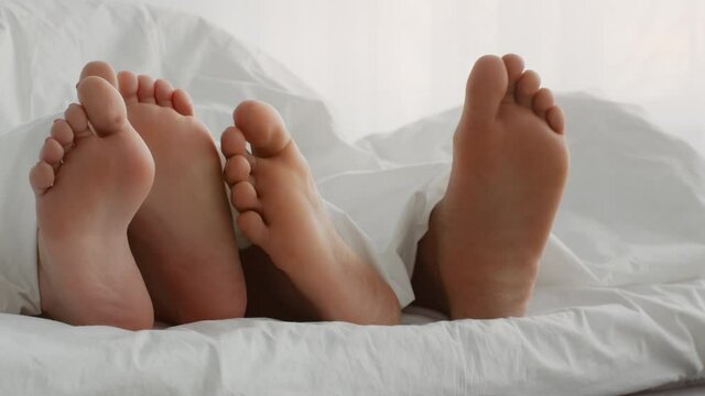 Young couple in bed lying in bed in morning, touching each other, close up shot of bare feet under blanket in bed