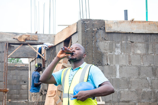 A Thirsty Male African Construction Worker Drinking From A Bottle On A Building Site And The Man Is Also Wearing A Blue Safety Helmet Known As Hardhat And Green Reflective Traffic Jacket